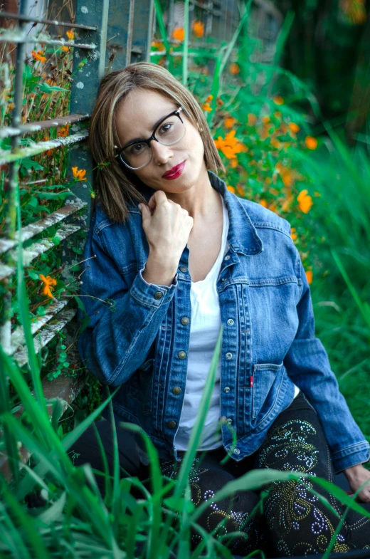 a woman in a jean jacket and glasses sitting against a fence