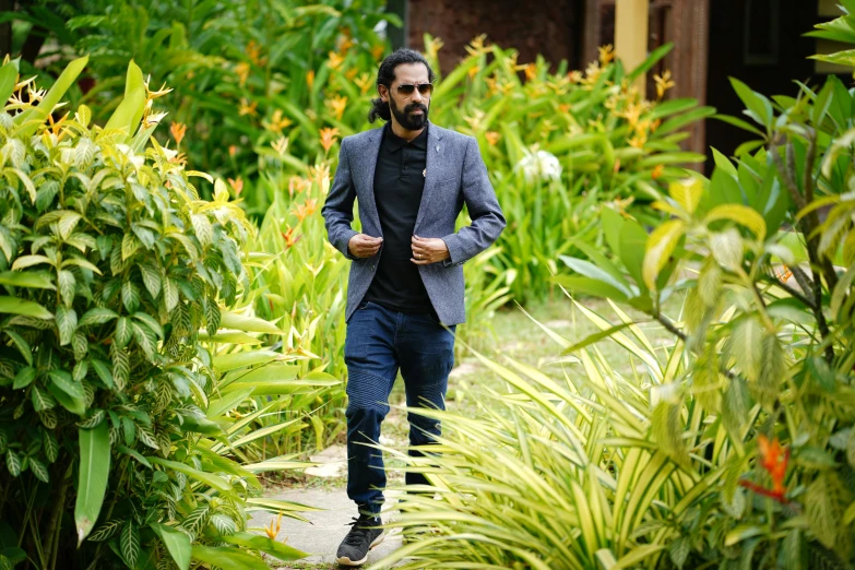 a man in a suit and sunglasses walks along the pathway through a garden