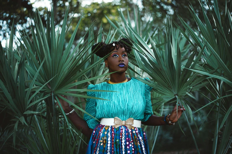 a black woman in a blue shirt and multi - colored skirt is standing among green plants