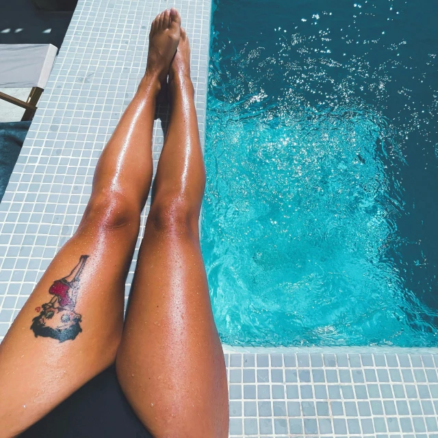 a woman sitting on the edge of a swimming pool, showing her bare legs and tattoos