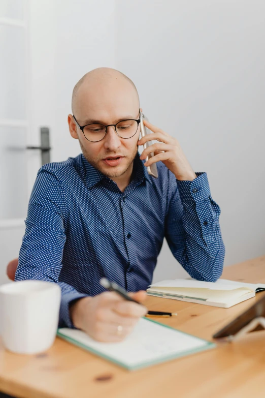 man sitting at desk with book and pen talking on cellphone