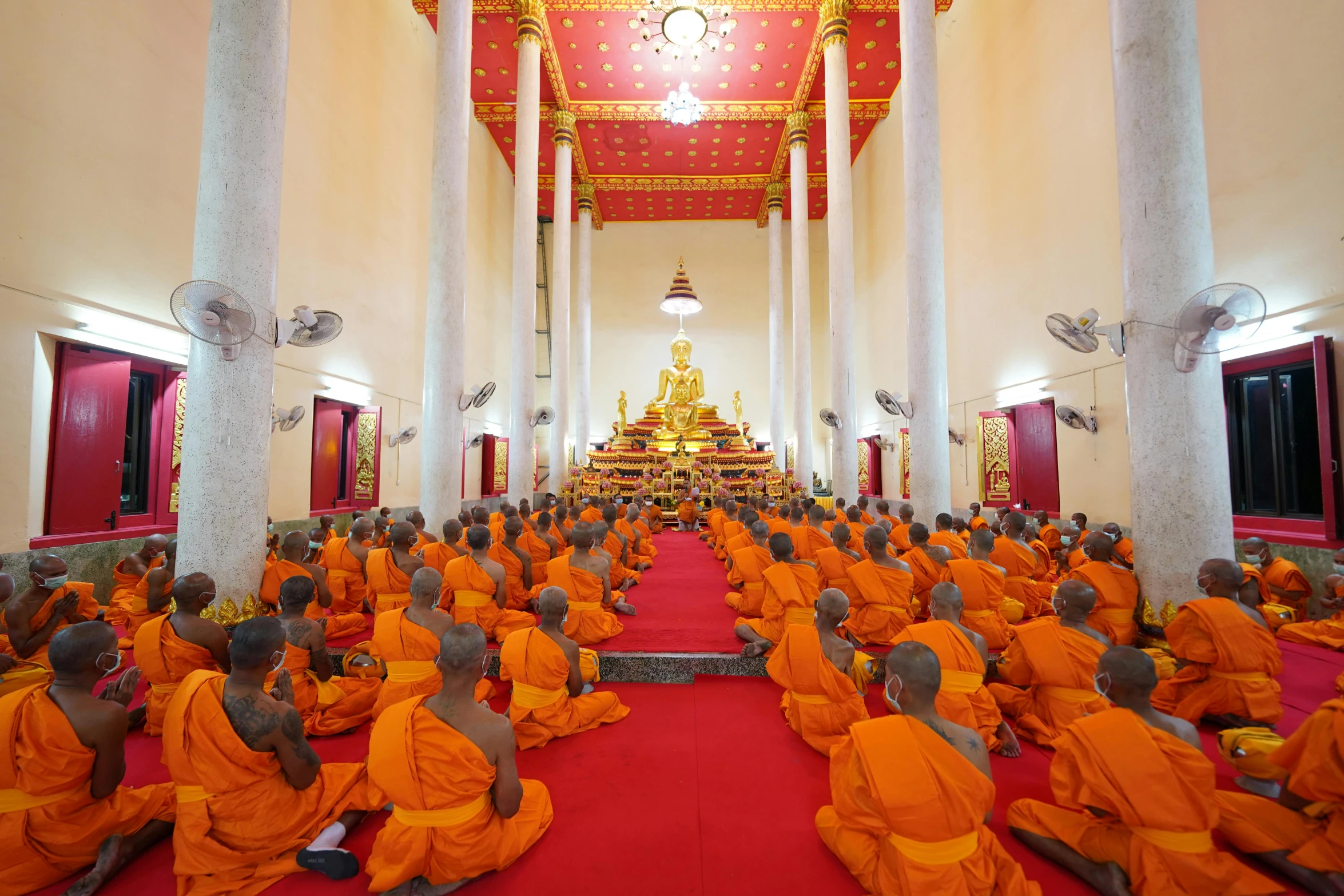 several monks sitting down in a long hall, wearing orange