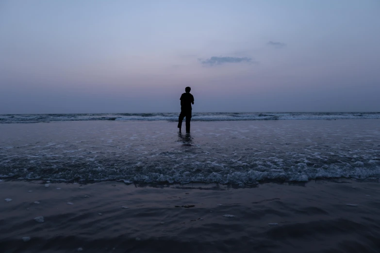 a lone surfer standing in shallow ocean water with purple and grey skies
