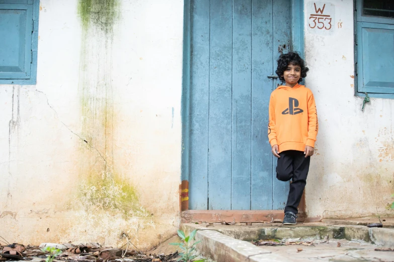 a child wearing a black and orange shirt leans against a building