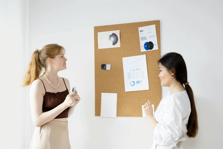 two women talking together while standing by a cork bulletin board