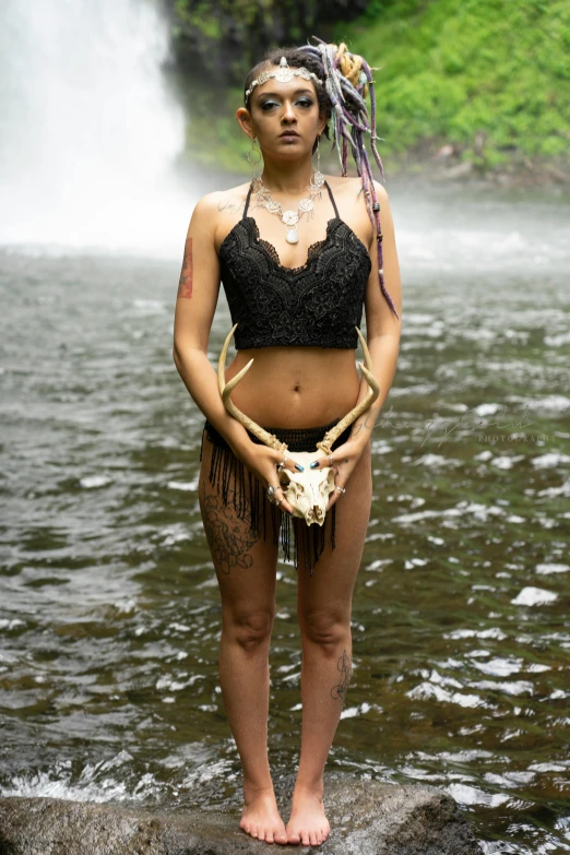 a person with weird jewelry and make up standing in the water near a waterfall