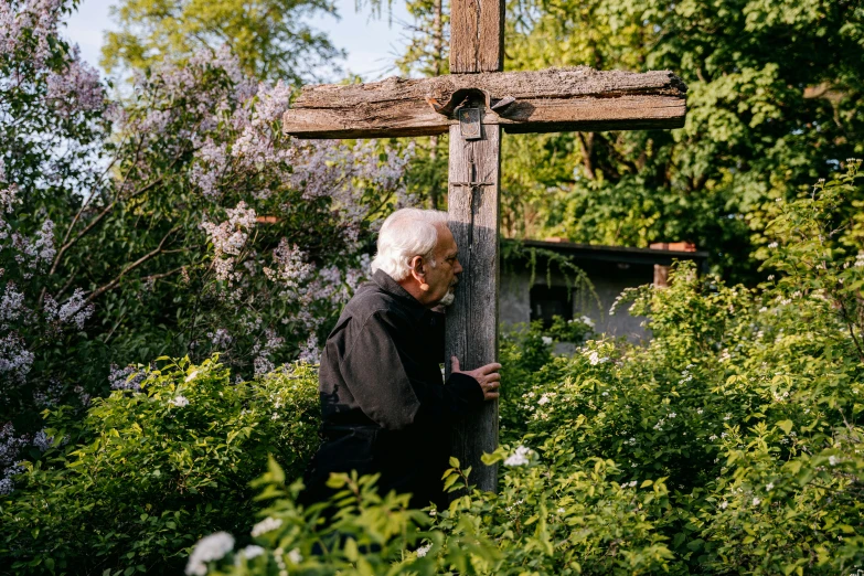 an elderly man in front of a wooden cross with lilacs