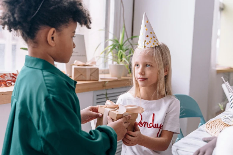 a little girl with a birthday hat is handing a gift to a boy