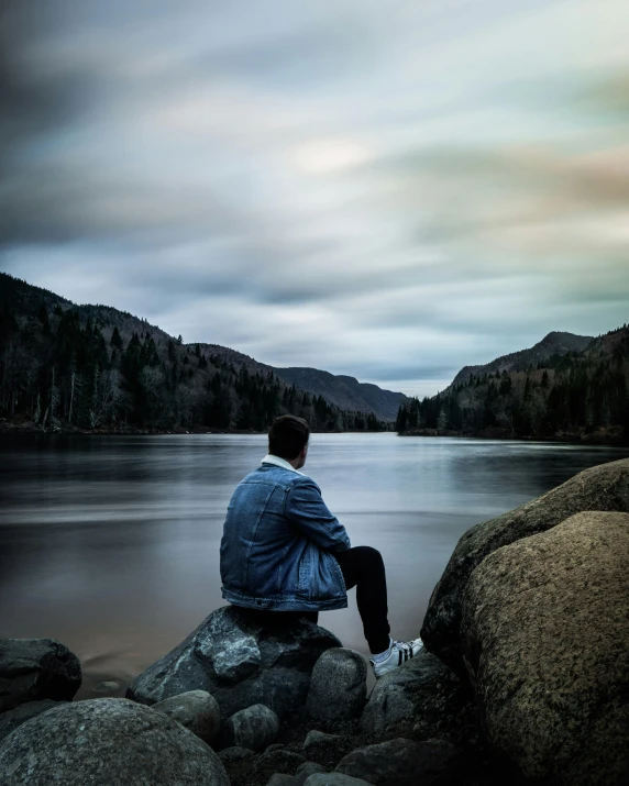 a man sits on rocks near water and looks over to the mountains