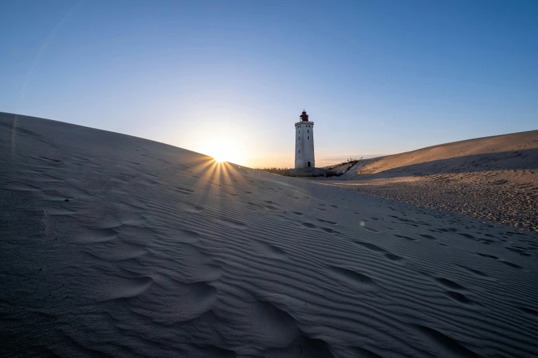 a light house sitting atop a sand dune with a sun setting behind it