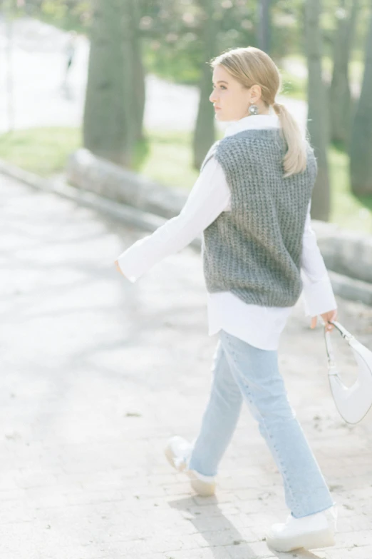 a woman wearing sweater vest and white sneakers holding a white bag