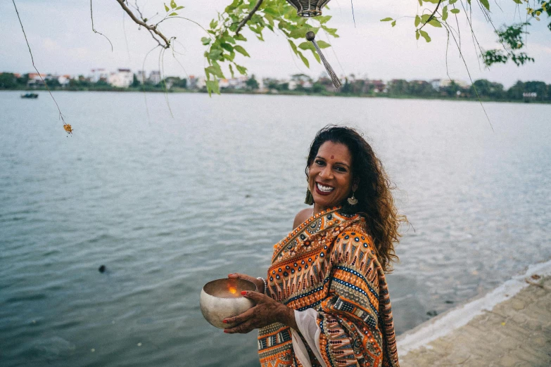 a woman holding a bowl under a tree by a lake