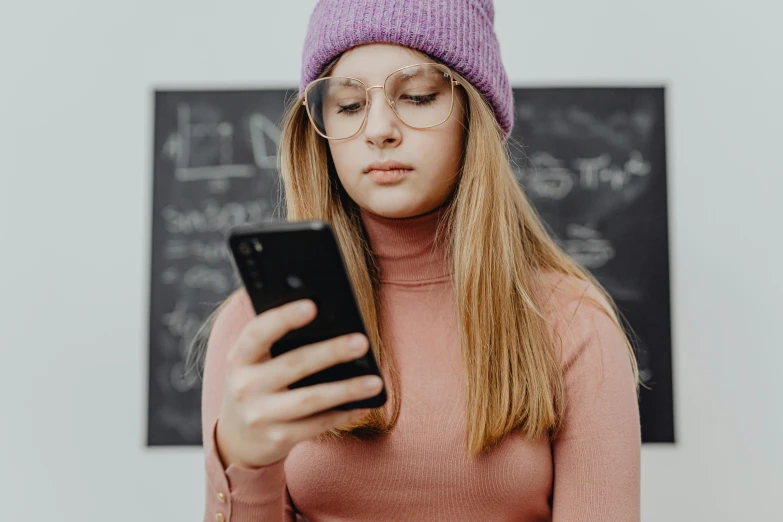 a woman with glasses, in a pink sweater and hat, checks her cell phone