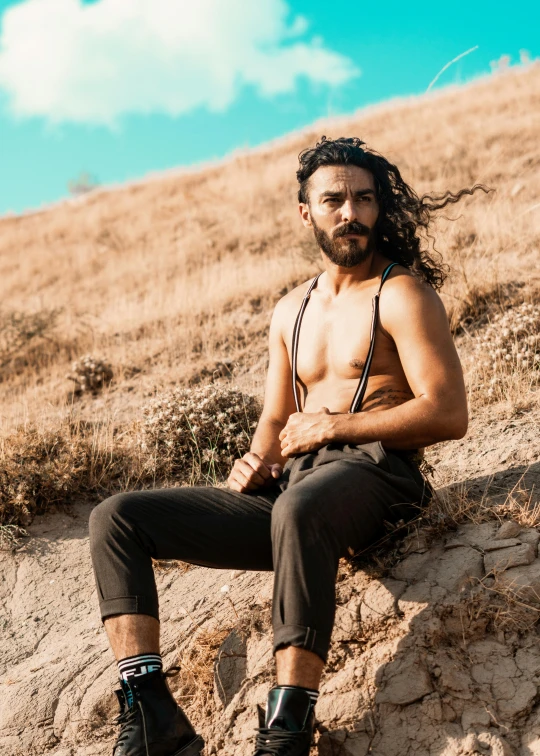 the shirtless man with his long hair hanging over his chest sits on a hill