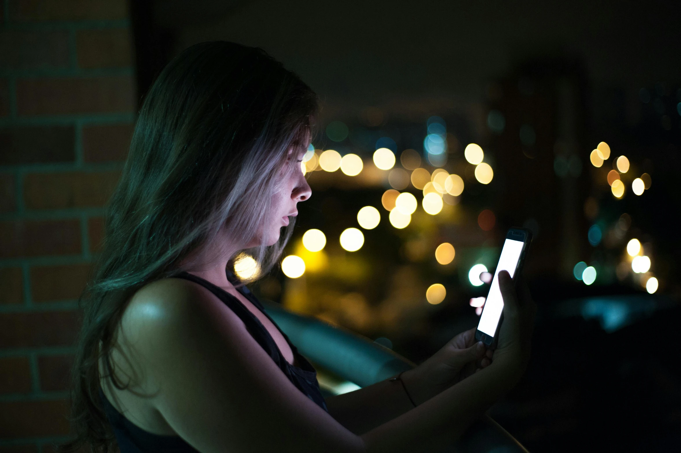 woman looking at a phone while sitting on the ledge in a dark area with some lights behind her