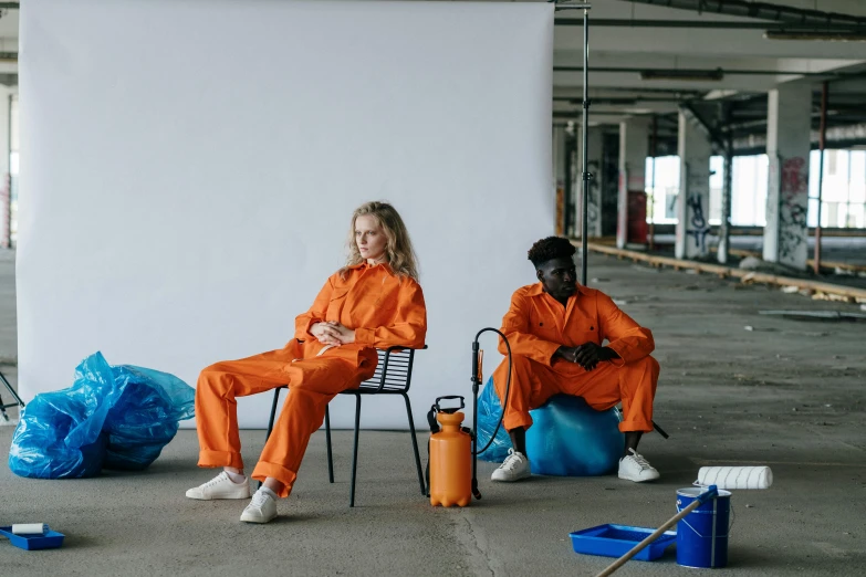 two women in orange jumpsuits sit on chairs next to garbage cans and other blue trashcans