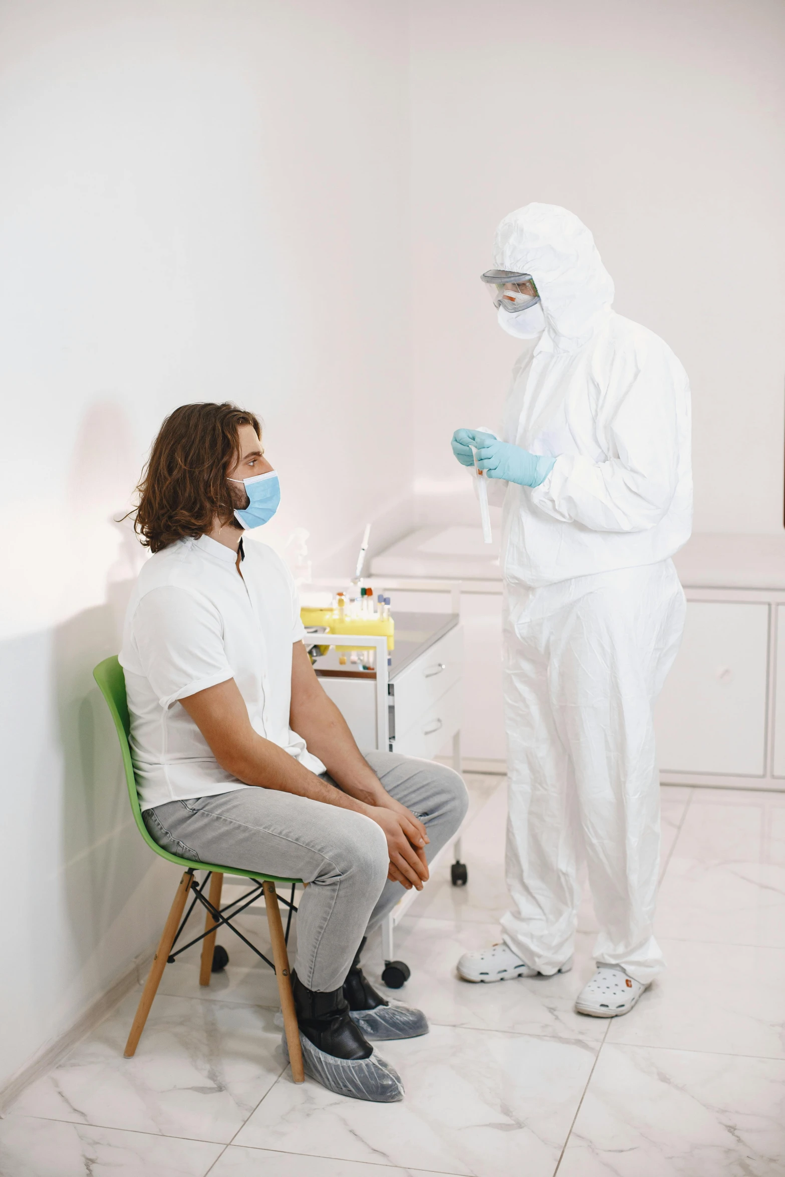 a person in a protective suit is sitting on a chair as a woman looks at him with a medical mask on