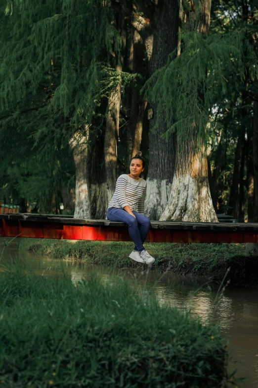 a young man sitting on the edge of a river next to a tree