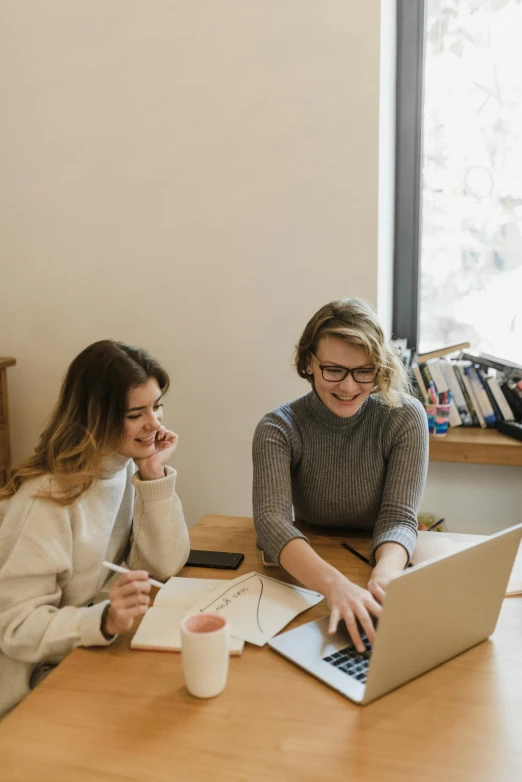 two women at a desk laughing over a notebook and laptop