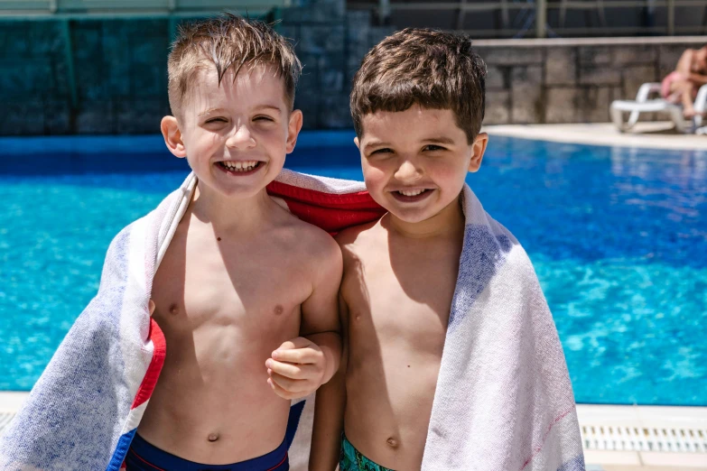 two boys in bathing suits holding up towels by a swimming pool