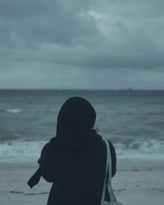 a person in black sitting on the beach with stormy clouds