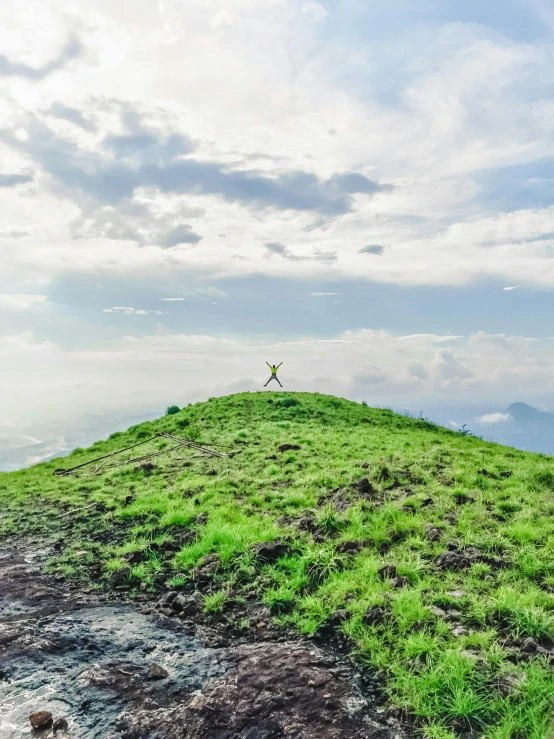 person jumping on top of green hill with cloudy sky