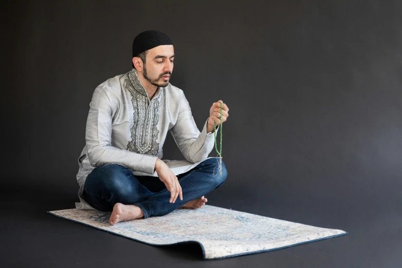 a man sits on top of a carpet, using a hook to tie beads