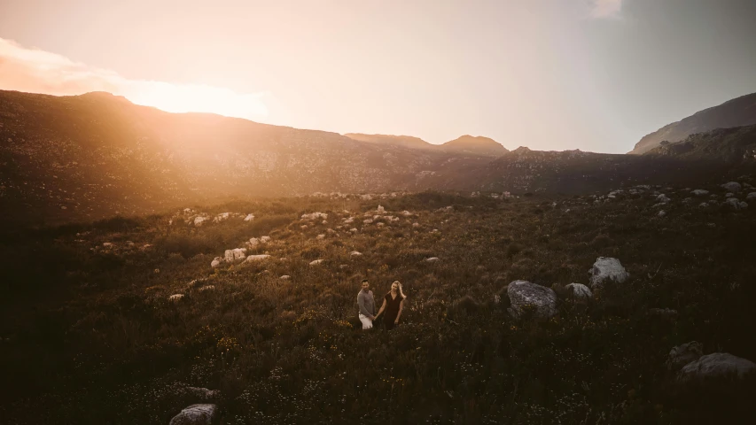 two people are sitting on a hill side while sheep graze