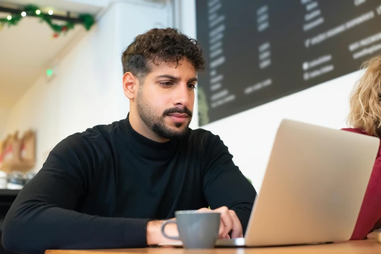 a man in black shirt sitting at a table using his laptop computer