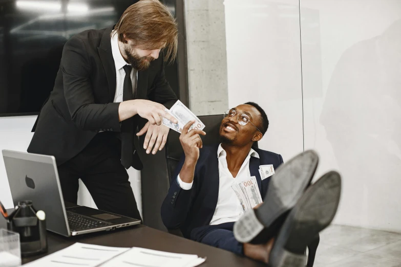 a man in a suit sits down at a desk and holds a paper near his face as another man sits in front of him