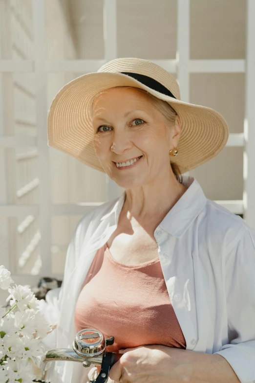a smiling woman wearing a large wide brimmed hat, holding a flower in her right hand