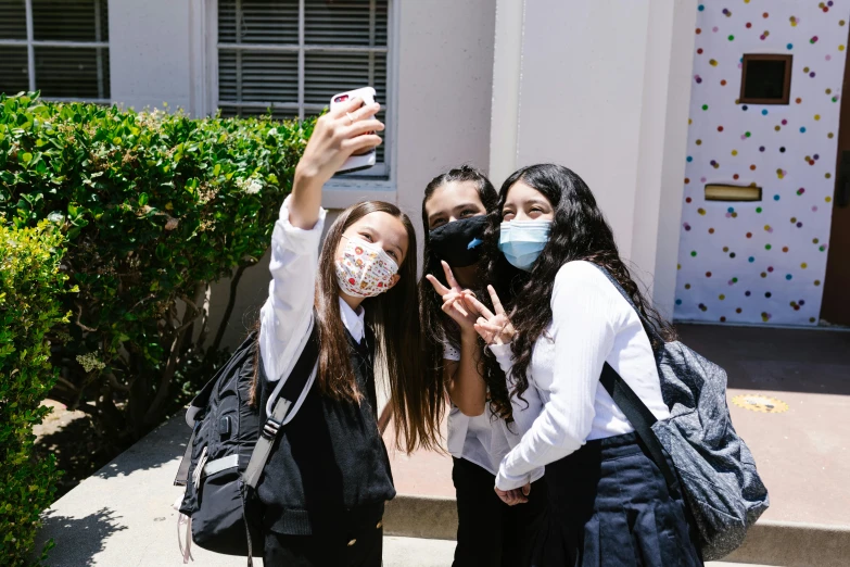 three young women standing outside a building holding their hands up and one woman with a face mask taking a selfie