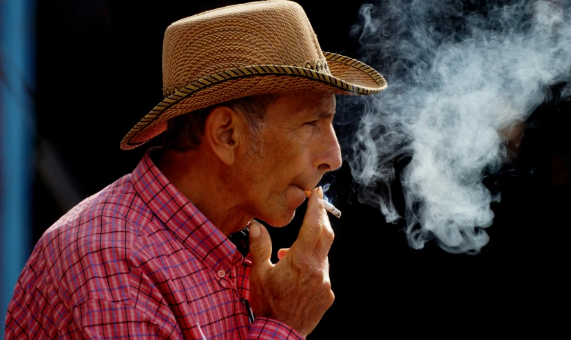 a man wearing a hat lights up a cigarette