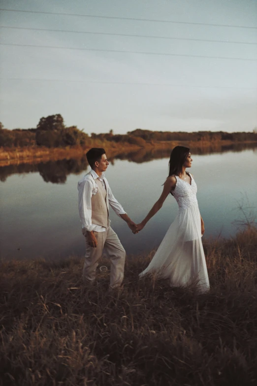two people dressed in wedding attire hold hands by a lake