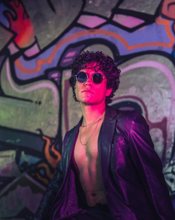 a man with glasses posing in front of a wall covered in graffiti