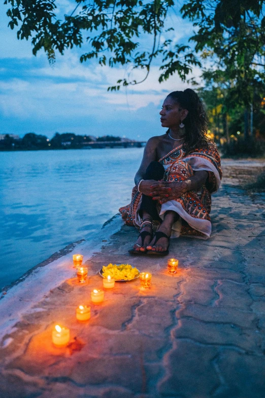 a woman sitting on a sidewalk next to water holding her hands in her pockets