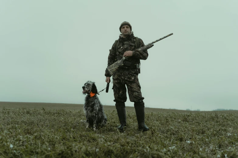 a man in full camo holds a gun and a dog in a field