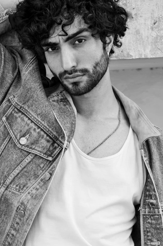 man in denim jacket and white tank top leaning against a wall