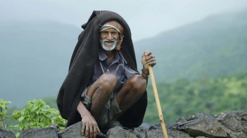 an elderly man with a long stick sits on top of a rock