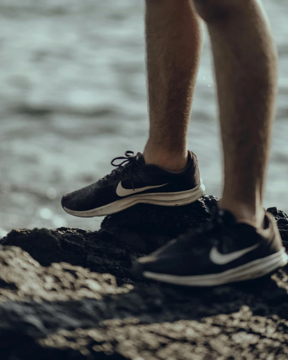 person's legs and running shoes on rock by the water