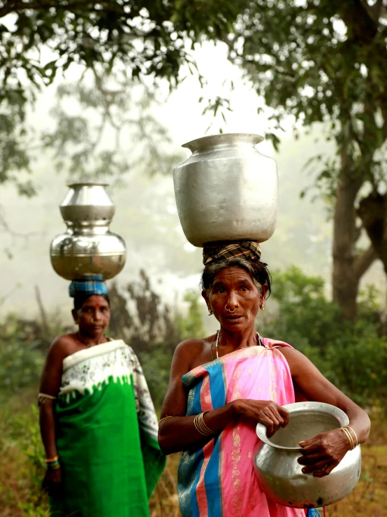 two women standing outside in a rural area holding bowls on their heads