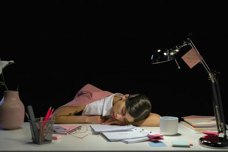 the woman lies down on a desk near a lamp, cup, and paper