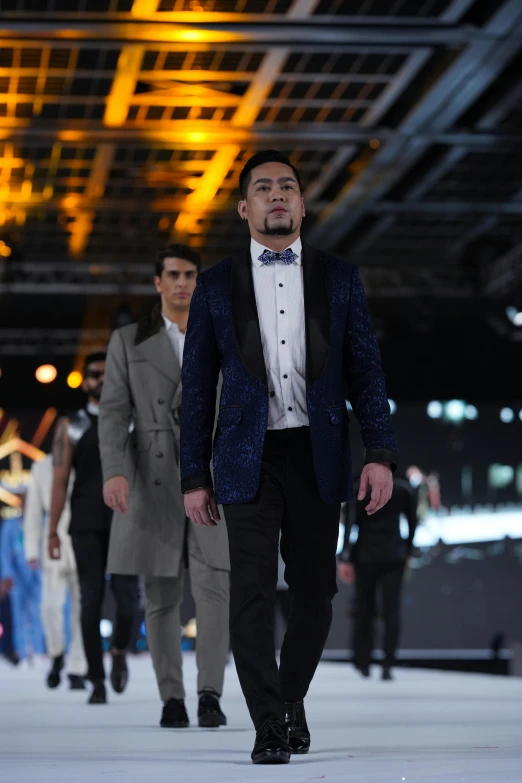 a man in a tuxedo walks on a runway with other men in suit