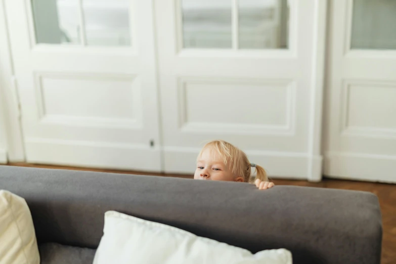 the young child has her eyes closed as she hides behind the gray couch