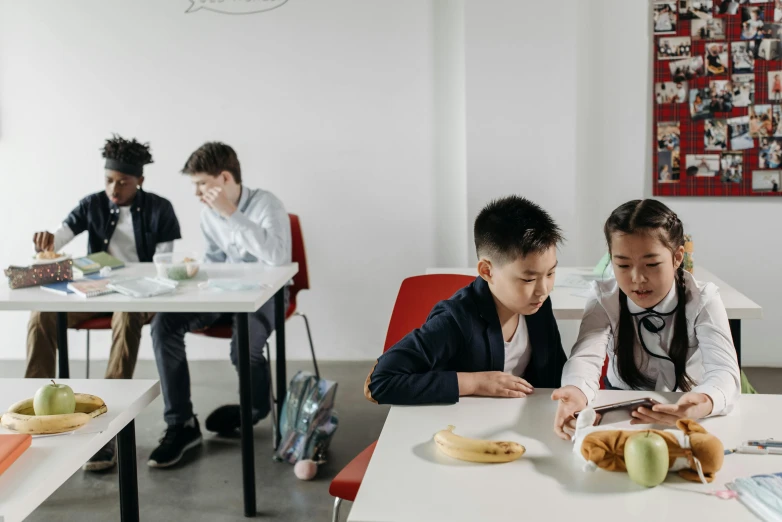 two children sit at a white table while one of them is reading and another child sits on a red chair