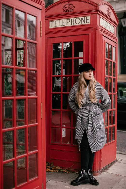 a woman in an unusual looking hat and coat stands next to a telephone booth