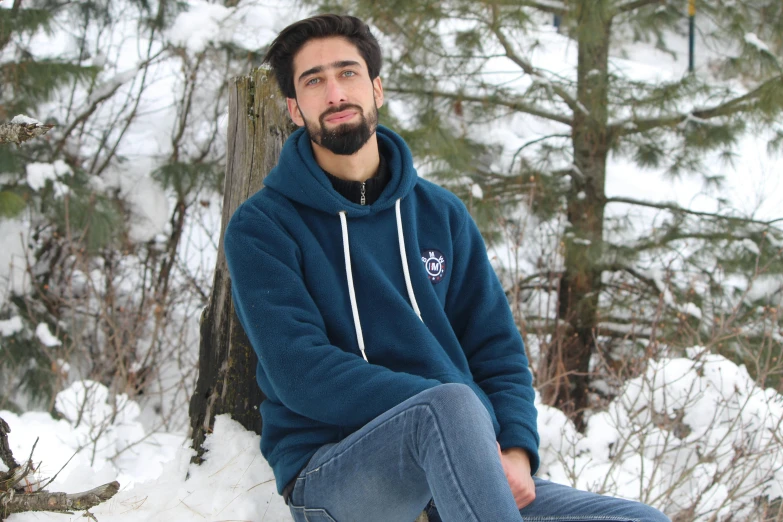 a bearded man in blue sits on a log next to a snow covered forest