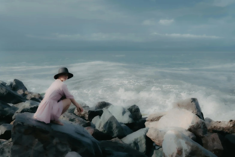 a woman in a hat and dress sits on rocks at the ocean