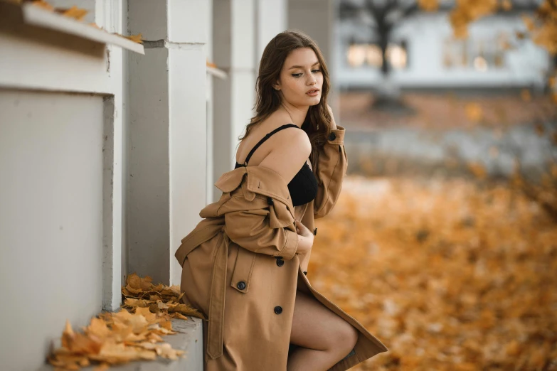 a woman wearing a trench coat leans against a pillar in the fall leaves