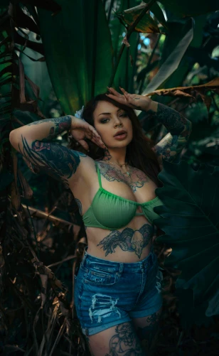 tattooed young woman in blue denim shorts leaning against leaves